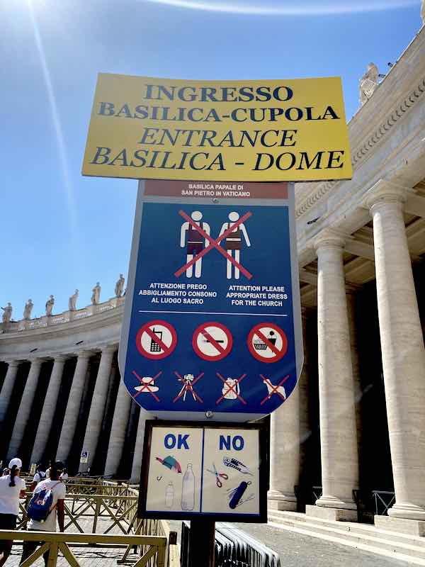 Sign in front of St Peter basilica with dress code for entry. Drawing of people in shorts and revealing clothing crossed out in red on blue background