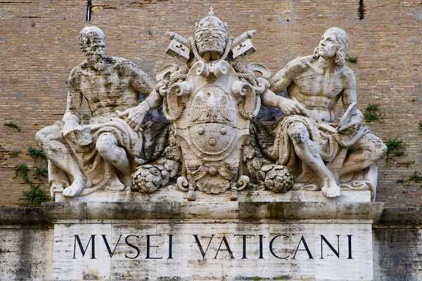 Sculpture at the entrance of Vatican Museum with two human figures, Papal Tiara and writing: Musei Vaticani